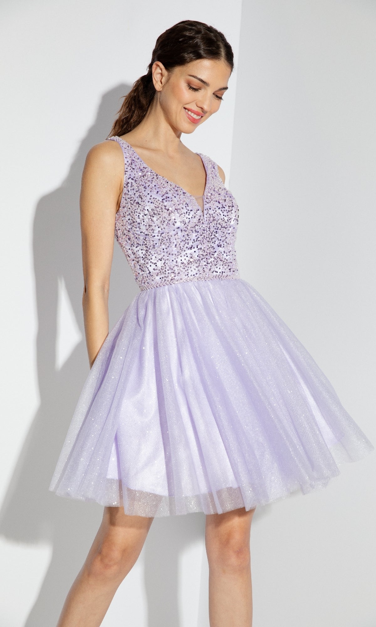 A woman in the Eureka 9926 Glitter-Bodice Short A-Line Hoco Dress, featuring a sleeveless sequin bodice and flared light purple tulle skirt, stands against a white background.